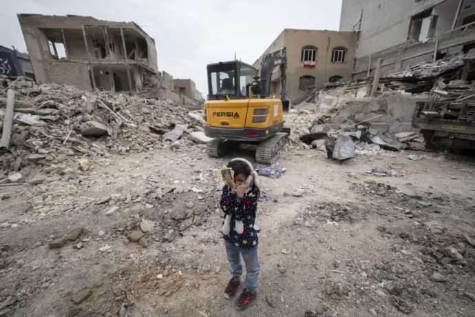 A young Iranian girl closes her eyes near a loader working on the ruins of buildings destroyed during a military campaign in Tehran on March 15, 2026.